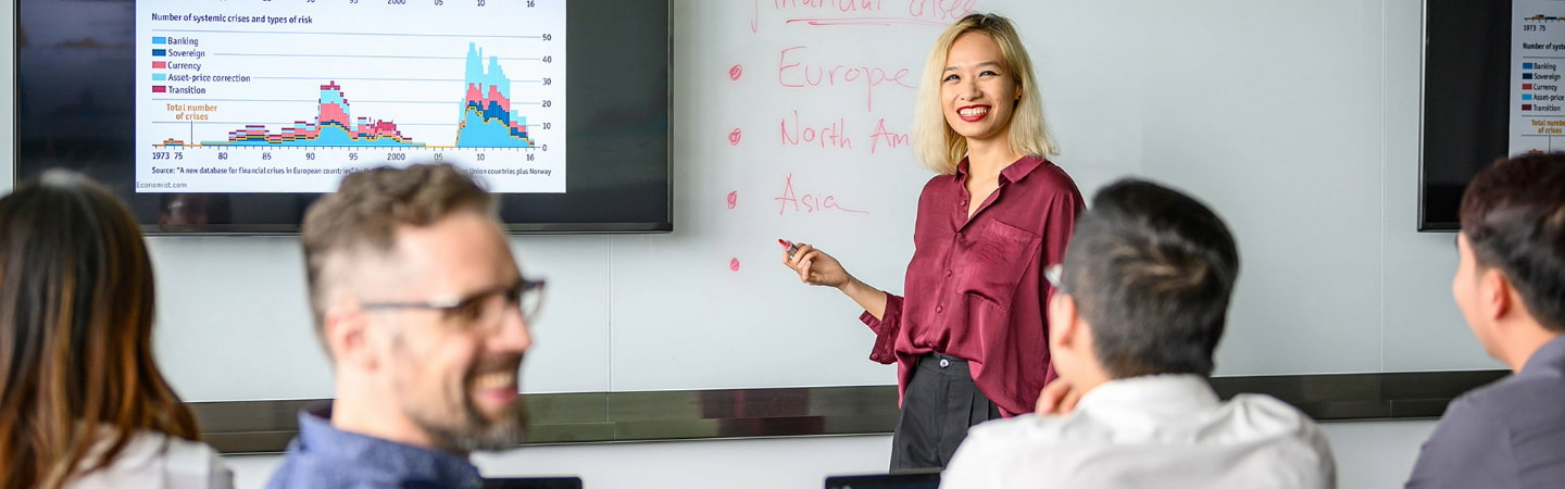 postgraduate group in class room, engaged in a discussion with visual data on a large screen and notes on a whiteboard.