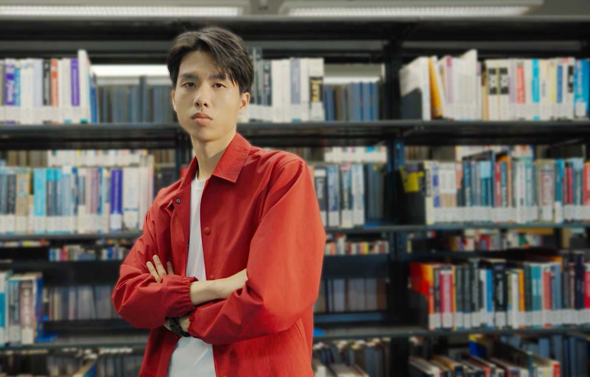 Tran Lam Nam Bao, winner of the 25th anniversary logo design competition, wearing a red shirt over a white t-shirt with his arm crossed, standing in the SGS library with the book stacks as a background. 