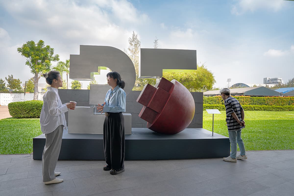 The image features an outdoor sculpture commemorating RMIT Vietnam's 25th anniversary, with three people standing nearby on a paved area surrounded by greenery.